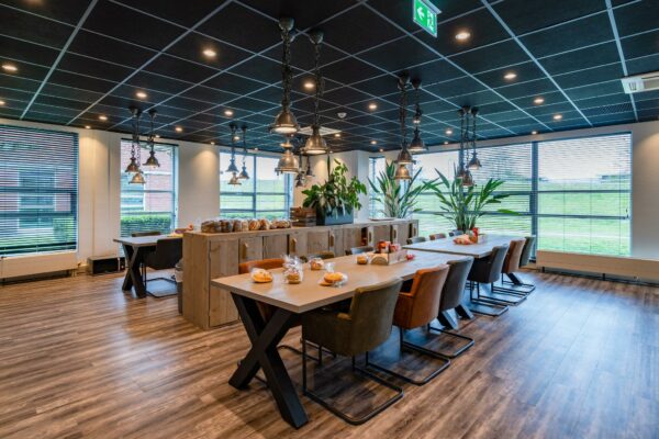 Moderne kantineruimte aan de 1e Barendrechtseweg met lange eettafels, hanglampen en verpakte broodjes.